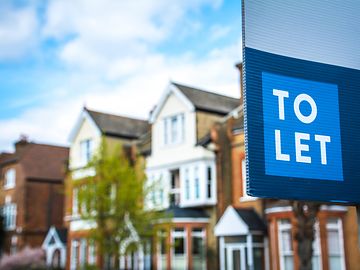 A blue to let sign, displayed in front of a row of read brick houses with white windows.