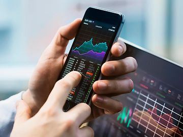 Finger pointing to phone on financial stocks app. Computer screen with financial stocks displayed behind.