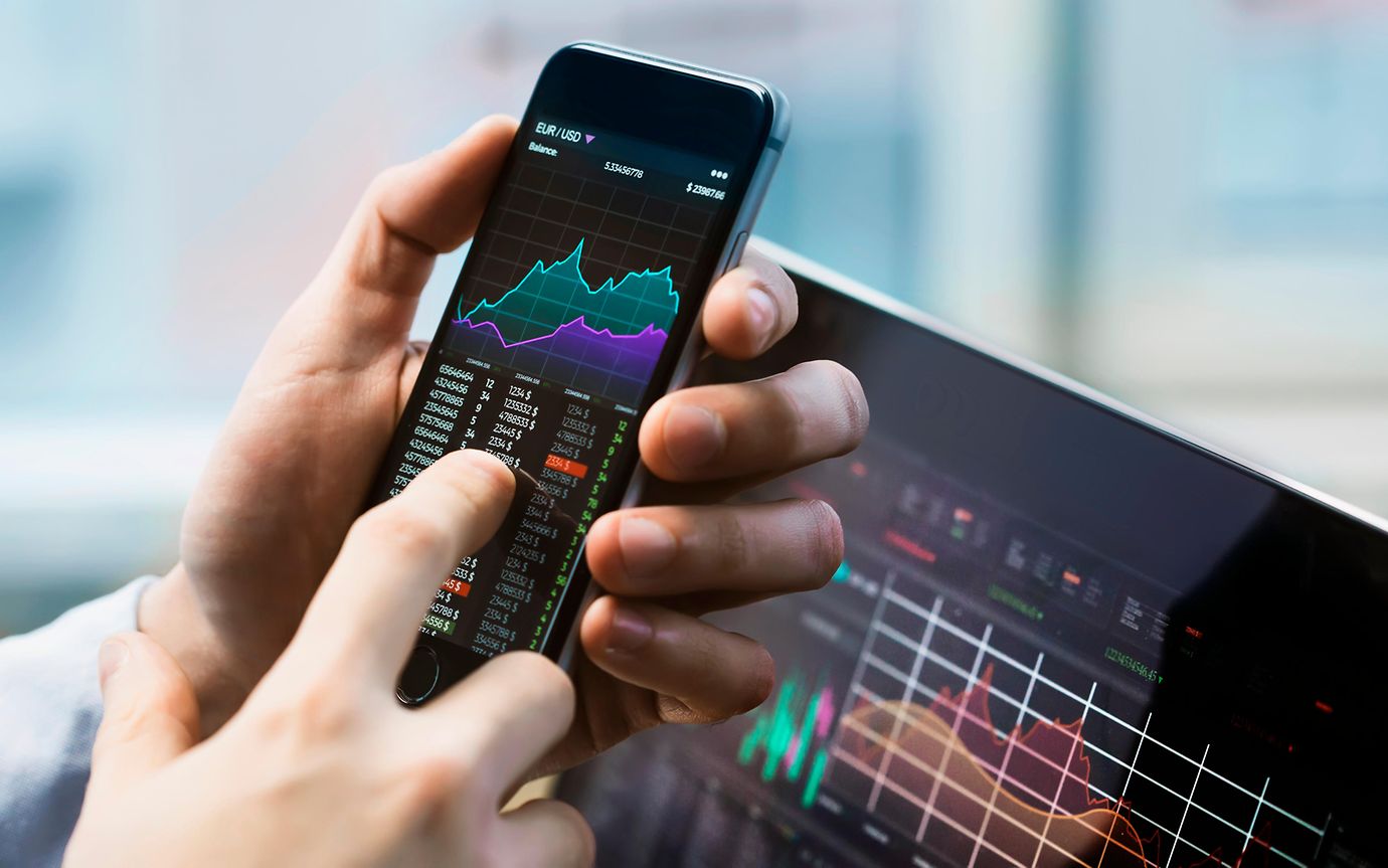 Finger pointing to phone on financial stocks app. Computer screen with financial stocks displayed behind.