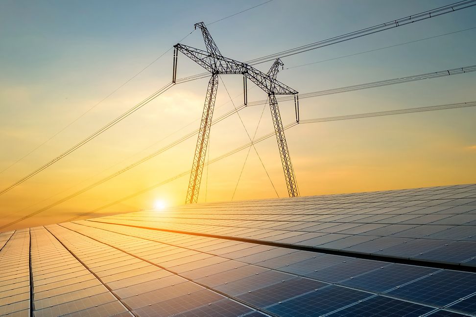 At sunrise, a steel high-voltage pylon with three strands of cable appears above a solar panel installation.