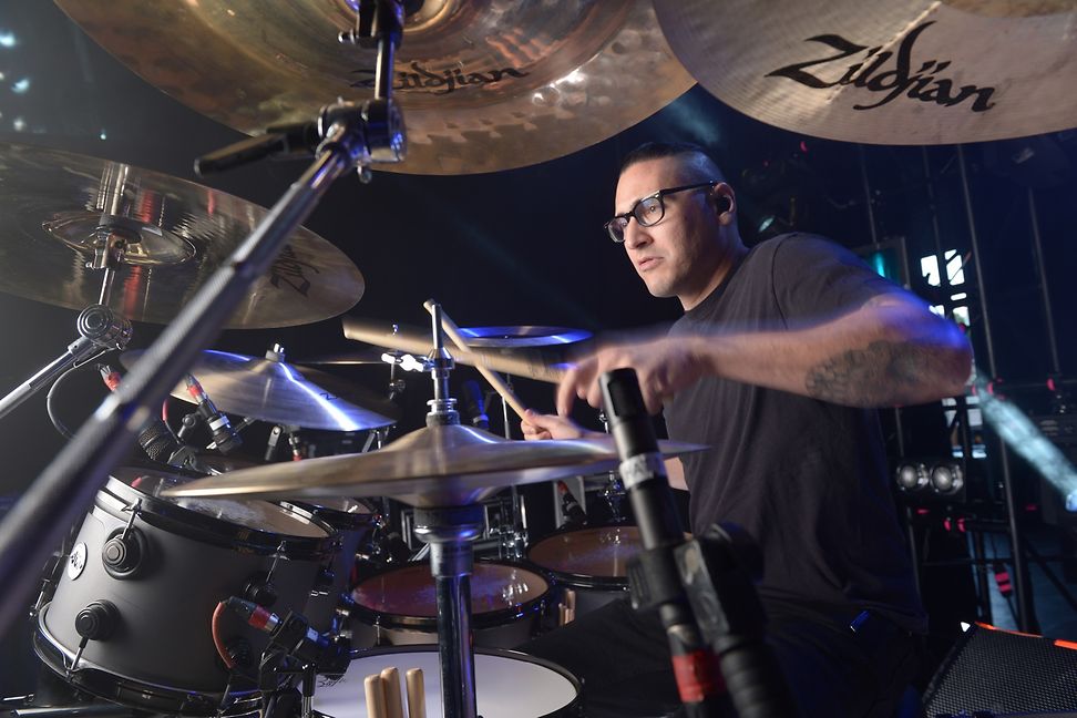 A drummer is playing on a large drum kit on a dark stage. Large cymbals can be seen in the foreground.