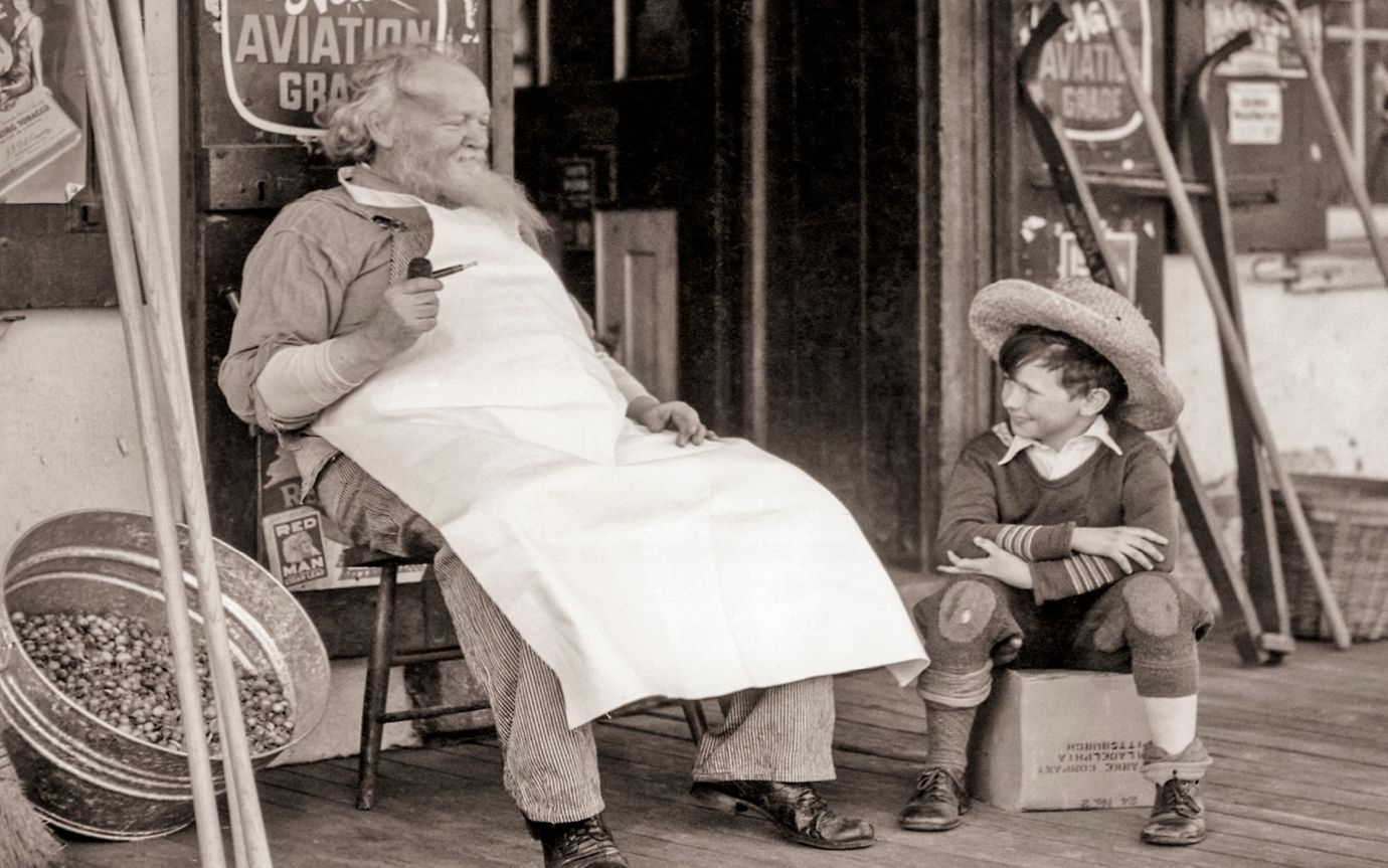 An elderly man in an apron sits on a chair in front of an old grocery store, next to a child perched on a crate.