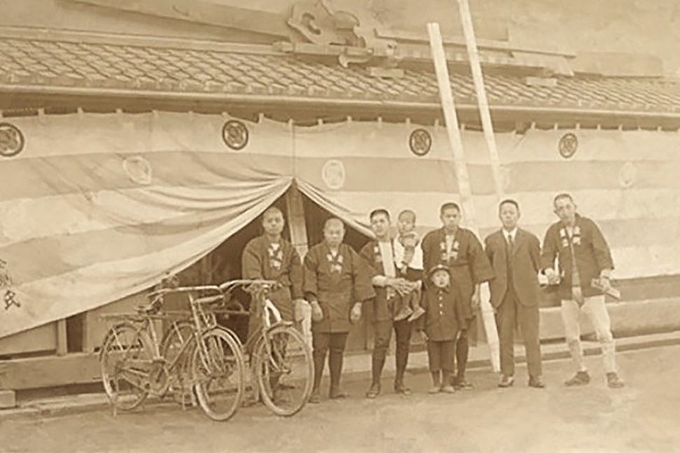 In an old sepia-coloured picture, a group of Asian men with two small children are standing in front of a building 